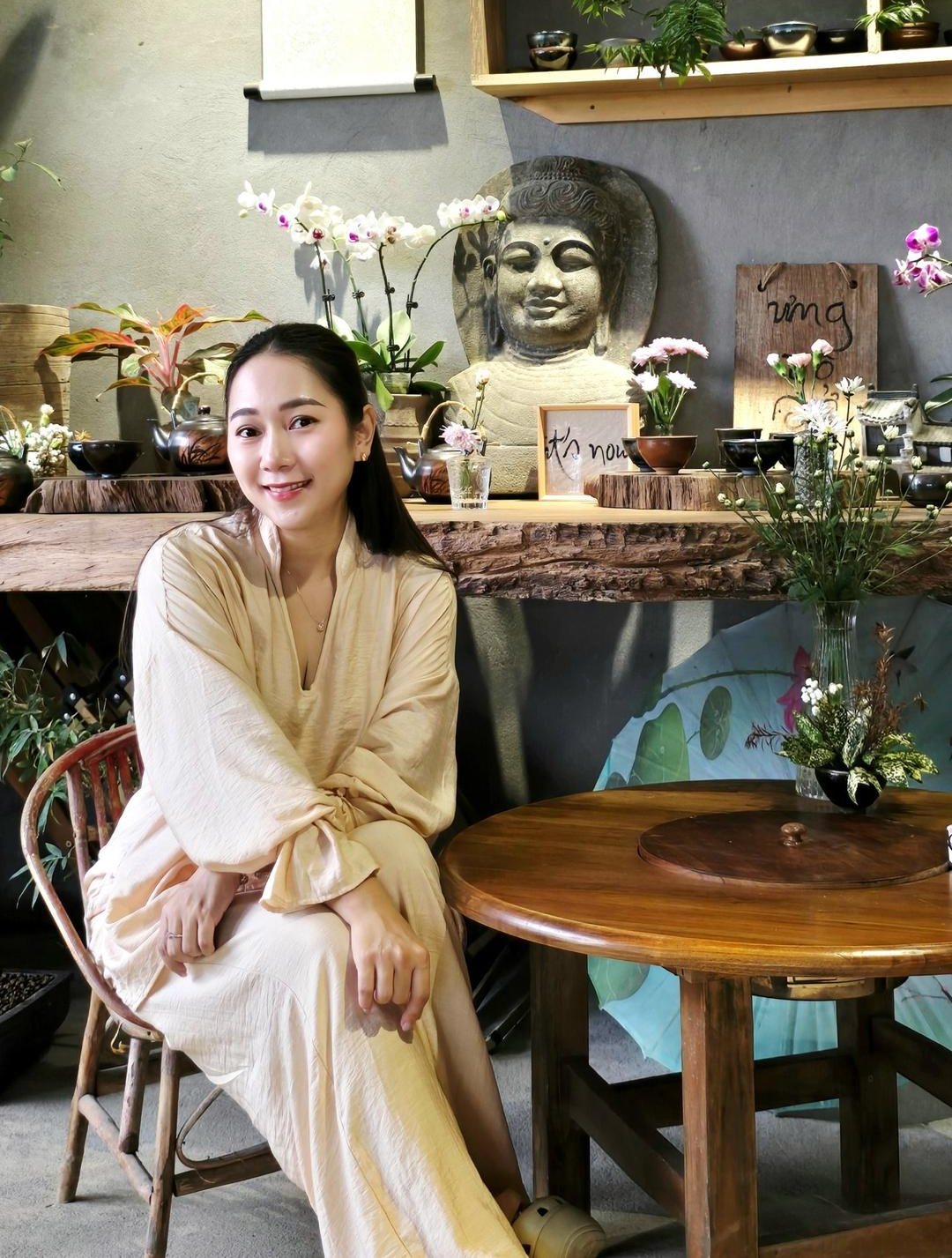 Grace mỉm cười trong bộ áo màu kem, ngồi cạnh bàn gỗ tròn trong phòng trà có tượng Phật đá, hoa phong lan và kệ gỗ đầy ấm chén