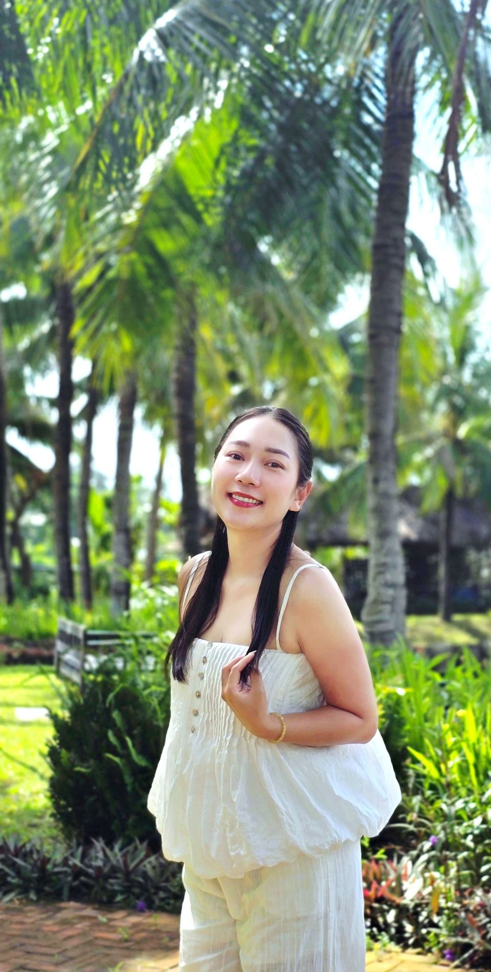 Grace mặc áo hai dây màu trắng, một tay chạm vào lọn tóc dài, mỉm cười giữa hàng dừa xanh