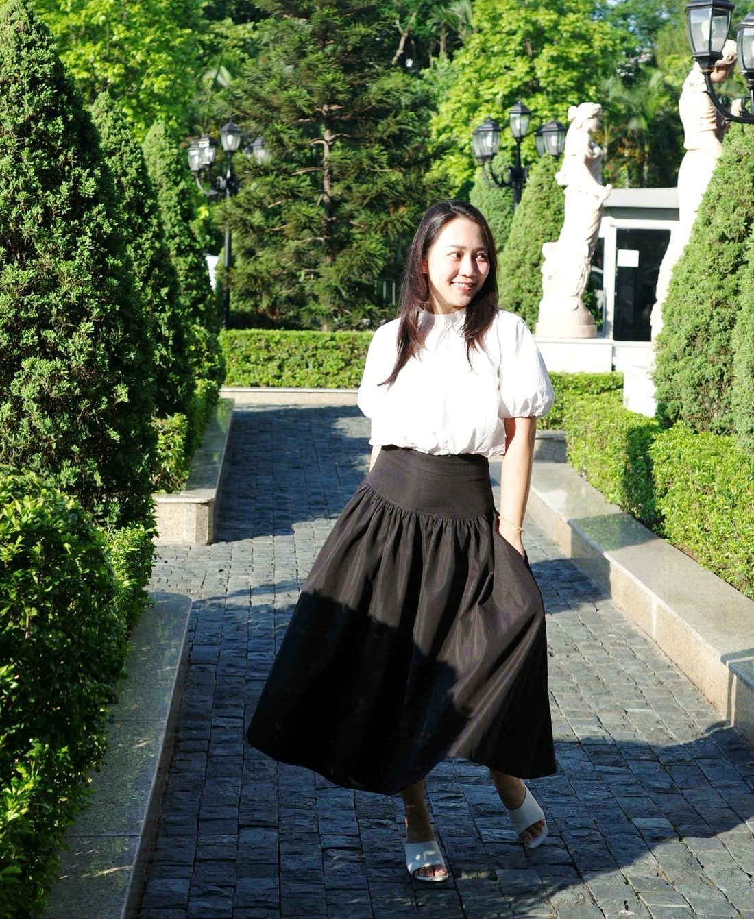 Grace mặc áo trắng tay bồng và chân váy đen xoè, bước nhẹ trên lối đá giữa khu vườn cắt tỉa gọn, quay sang một bên và mỉm cười