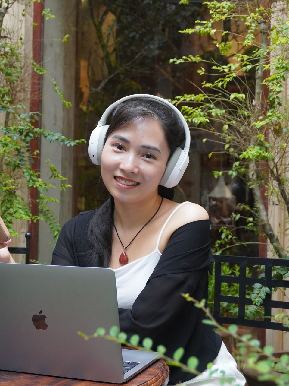 Annie đội tai nghe trắng, ngồi bên chiếc bàn gỗ trong khu vườn nhỏ với laptop và điện thoại, mỉm cười nhìn thẳng về phía bạn