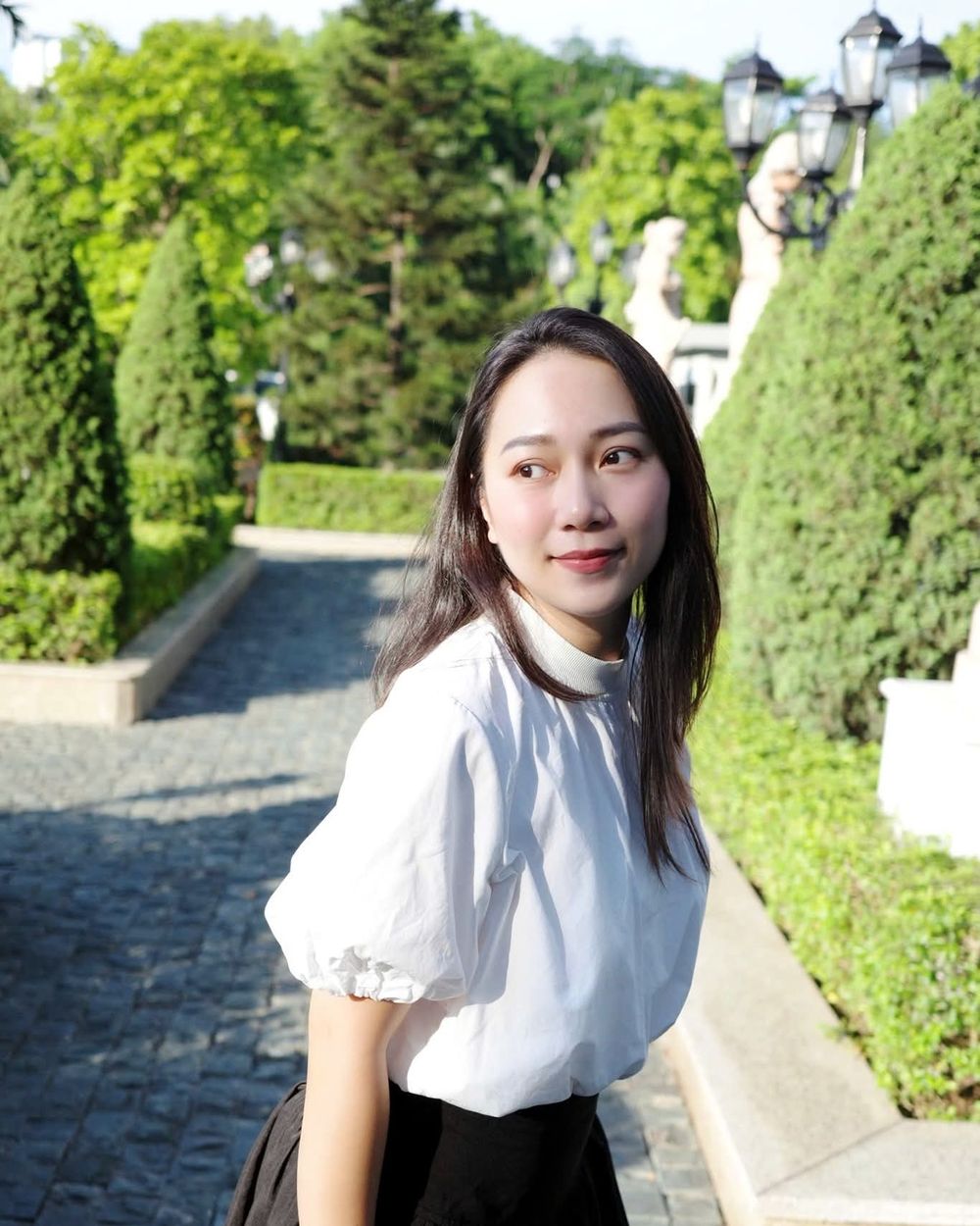 Grace trong chiếc áo trắng tay phồng, đứng giữa lối đi vườn cây xanh, quay đầu nhìn qua vai với ánh mắt dịu