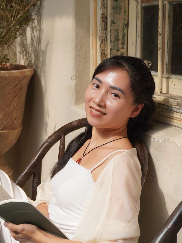 Annie Phan ngồi bên cửa sổ, mỉm cười, tay cầm một cuốn sách