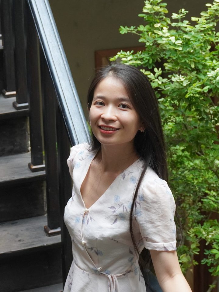 Thuỷ Nguyễn đứng trên cầu thang gỗ, mỉm cười, phía sau là cây xanh