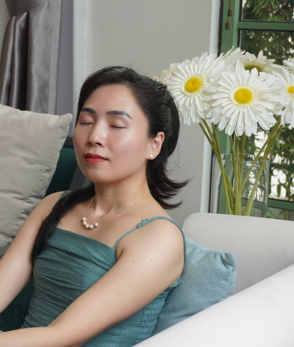 Annie ngồi tựa lưng trên ghế sofa, mắt nhắm lại và thở nhẹ, bên cạnh là bình hoa cúc trắng