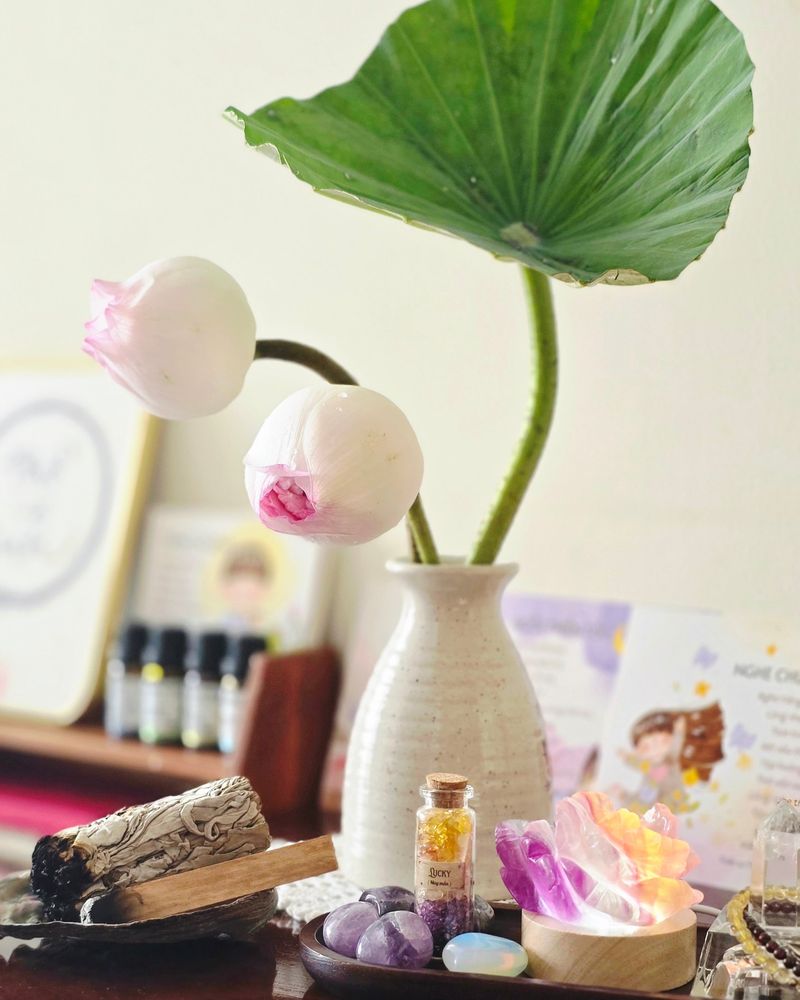 Chiếc bình gốm trắng cắm lá sen và hai nụ sen, bên cạnh những lọ tinh dầu nhỏ và viên đá tím