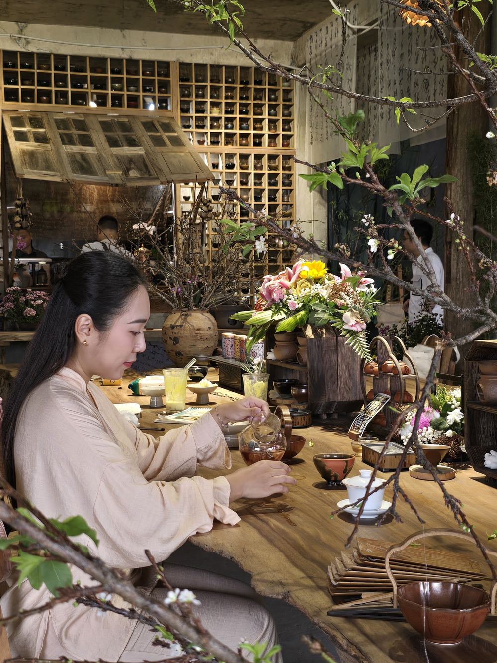 Grace ngồi bên chiếc bàn gỗ dài trong một quán trà đầy hoa và cành cây, tay đang rót trà