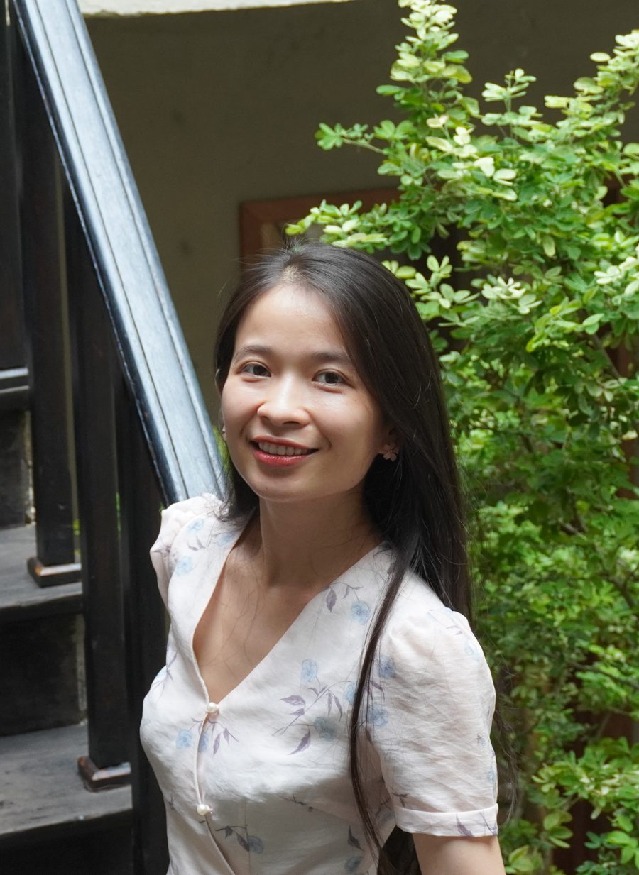 Thuỷ đứng bên cầu thang gỗ tối giữa những tán cây xanh, mặc váy hoa nhạt, mỉm cười nhìn về phía bạn