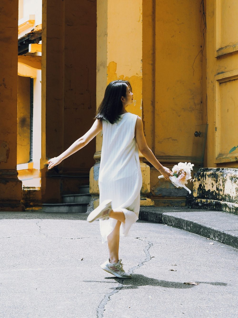 Một người phụ nữ mặc váy trắng xoay người nhẹ nhàng giữa khoảng sân vàng, một tay dang rộng, tay kia cầm bó hoa